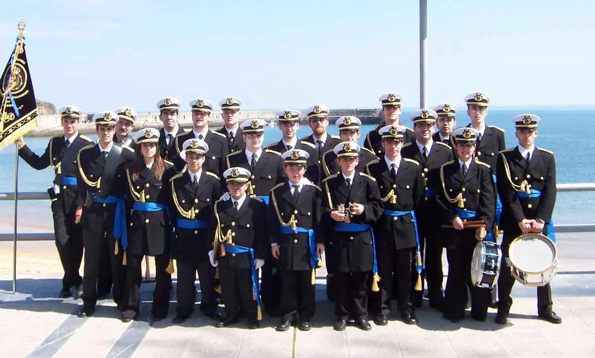 Foto de grupo de la agrupación junto al mar en Luanco, con niños y adultos en uniforme azul y blanco.