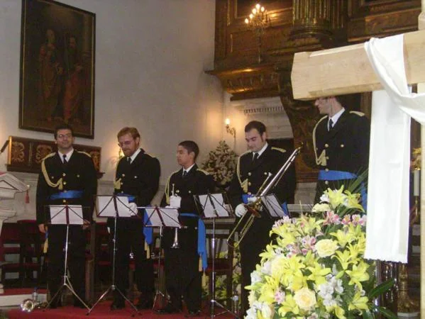 El grupo de metales de la agrupación toca de pie con sus atriles en el interior de una iglesia, junto a una cruz vestida.