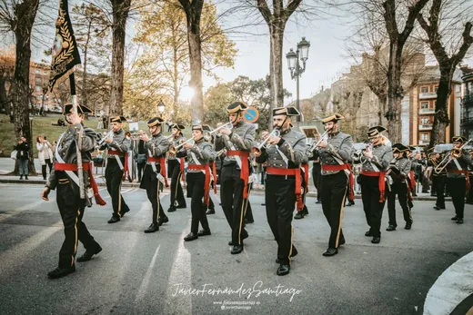 La agrupación desfila tocando por un paseo arbolado, con el banderín al frente.